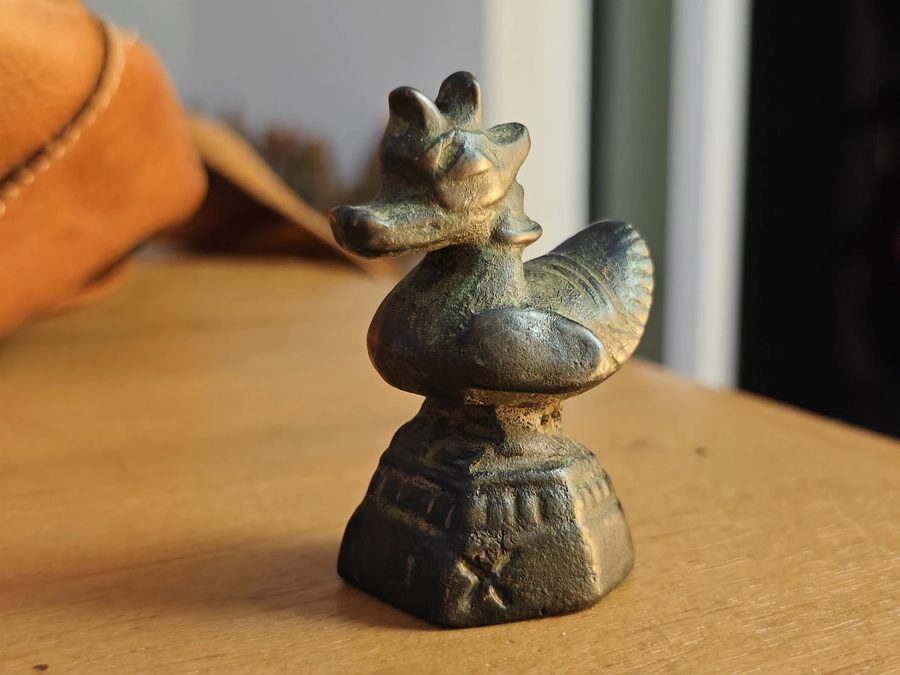 Photo: a small bronze swan, a hongsa (หงส์), sitting on a stepped pedestal on a wooden table in low, warm sunlight. The bird has a crest of three rounded points on its head, closed eyes, a long flat beak that curls up at the tip, a ring around its neck, a round chest and a fanned tail with carved lines. The metal is dark brown, gold where hands have rubbed it smooth, and green in the hollows. The pedestal has a band of upright marks and a cross scratched into its front. Out of focus behind it: a white wall, a dark window frame and a tan leather bag. Its shape is the shape of the old bronze bird weights of Burma and northern Thailand.